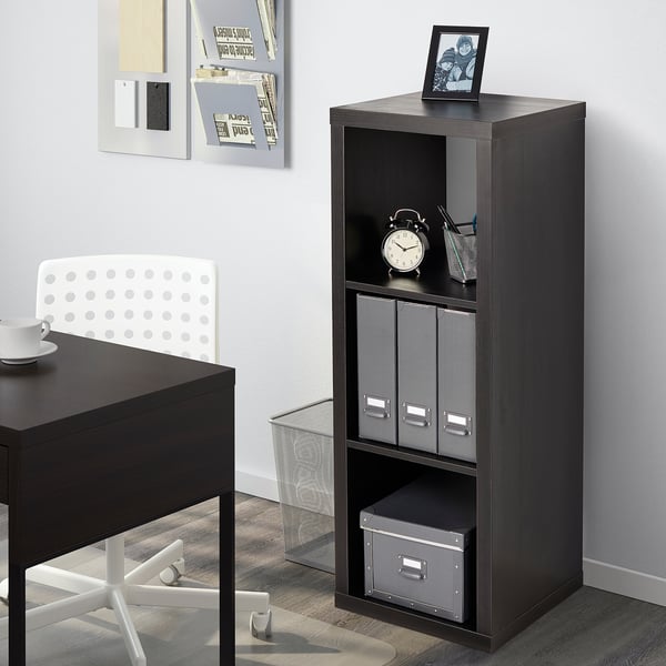 Das schwarze KALLAX Bücherregal steht neben einem Schreibtisch und hält eine Uhr, Schachteln und ein Foto. Die Regale sind modular aufgebaut und lassen die Gegenstände ordentlich verstauen.