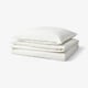 Clean white bedding stack on white background. Includes folded duvet and pillowcases in soft, washed cotton. Emphasises comfort and simplicity.