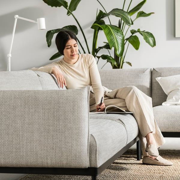 Comfortable person writing on couch, cream blouse and pants, large leafy plant behind, white lamp, modern grey sofa, cosy ambiance.