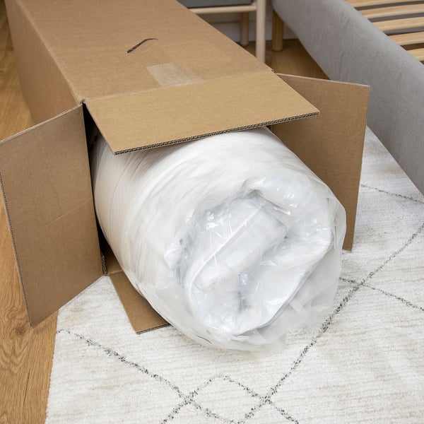 Cardboard box containing rolled, vacuum-packed mattress with cling film, placed on a rug.