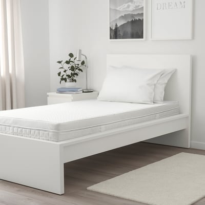 Minimalist bedroom: white ÅVABERG bed, rustic floor, wall art. Bed is small, with two pillows, plant, books, and lamp on bedside table.