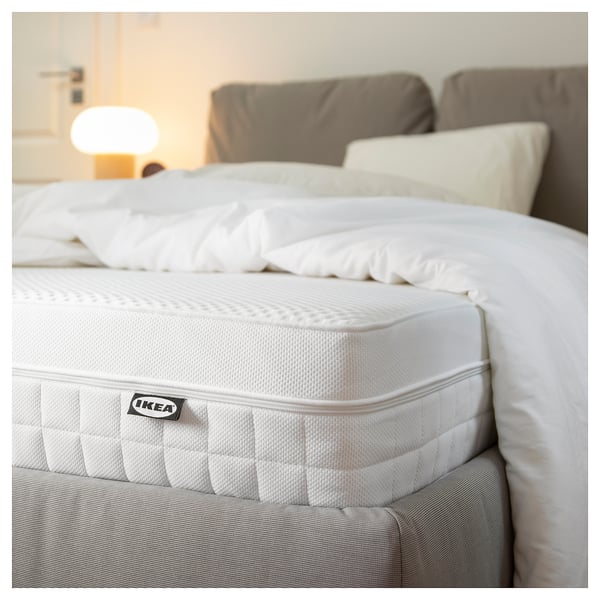 A corner of an IKEA ÅVABERG white mattress on a bed with white sheets and a grey bed frame, highlighting its foam texture and comfort zones.