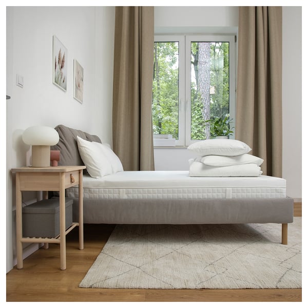 Modern bedroom with white mattress on grey bed, large window, side table with lamp, and rug under bed.