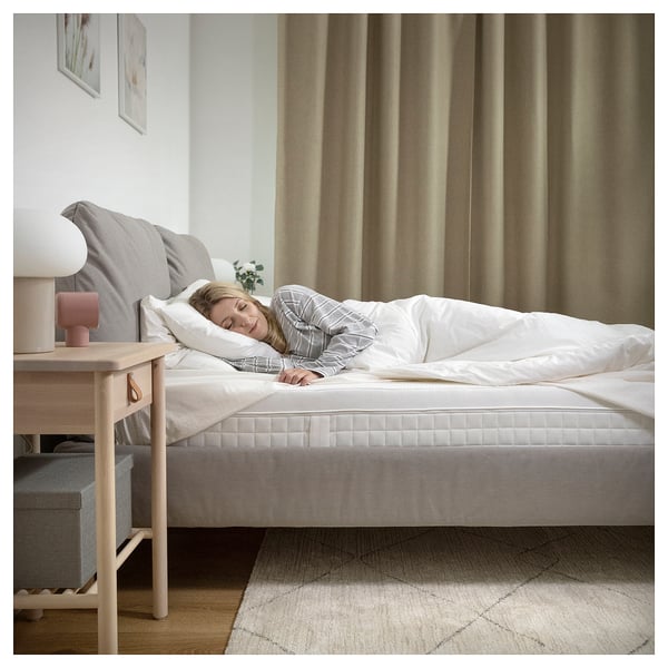 Cosy bedroom with ÅVABERG white mattress, side tables, lamp, and curtains. Person sleeping peacefully.