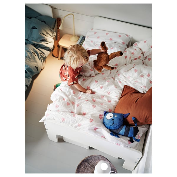 Kid playing on bed with heart print duvet cover, holding teddy bear, blue cat toy beside.