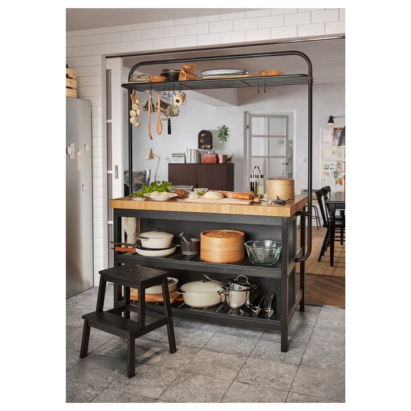 Modern kitchen with a wooden island table and black-step stool. Various pots, pans, and utensils organised on shelves and hanging racks.