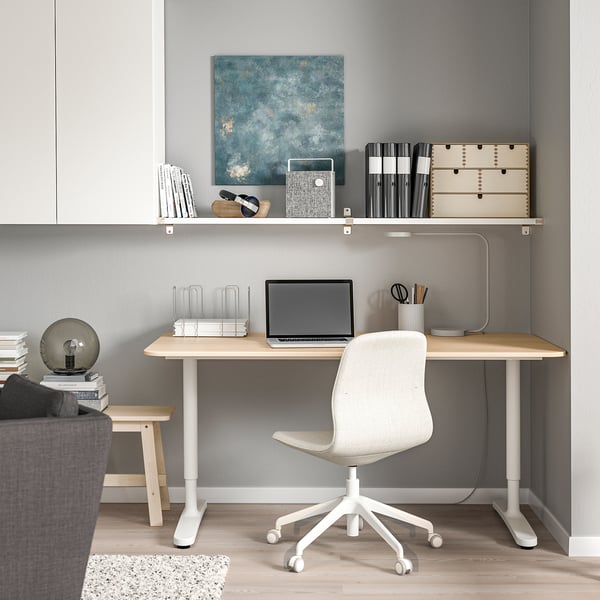 Modern home office with light wooden desk & white chair, laptop, books, & BERGSHULT wall shelf above. Grey walls.
