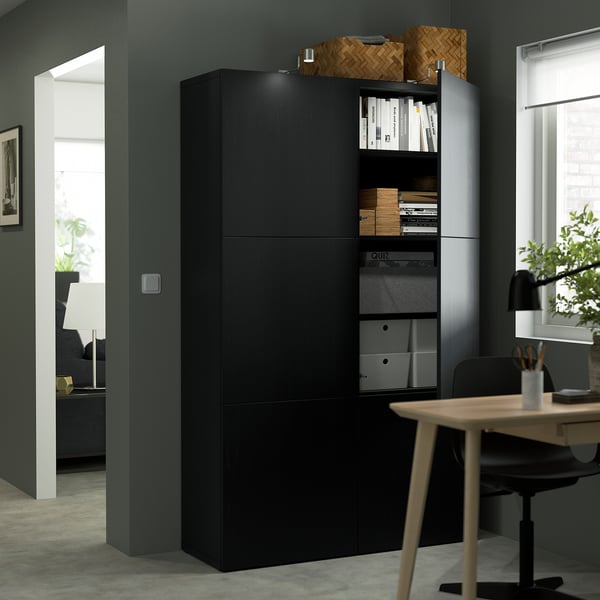 Modern home office setup with black BESTÅ storage cabinet featuring adjustable shelves, books, and a wooden desk with a chair.