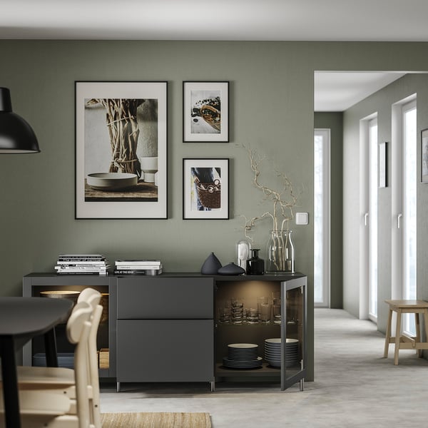 Modern dining room with dark grey BESTÅ sideboard, green walls, and minimalist decor. Include framed art, glassware, and stacked plates.