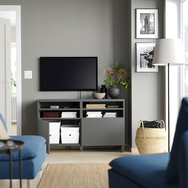 Modern living room with grey BESTÅ tv stand, flat-screen tv, blue sofa, woven basket, lamp, books, and black and white photos.