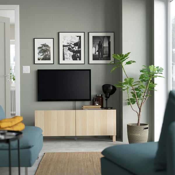 Modern living room with BESTÅ light wood tv bench, sage green walls, art, plant, teal couch & storage.