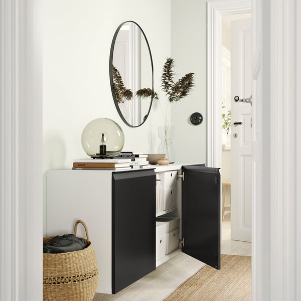 Modern entryway with white BESTÅ cabinet, black doors, round mirror, dried plants, wicker basket, and open door leading to a white room.