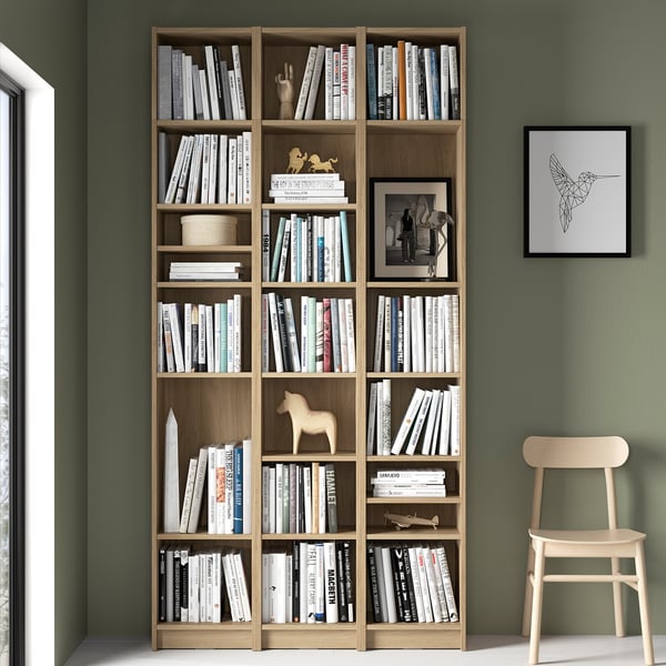 BILLY bookcase in natural wood, filled with books and decor, against green wall, taller than light wood chair.