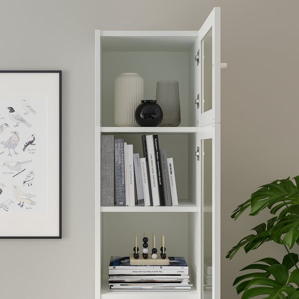 A white BILLY bookcase with glass doors books and decorative items. Adjustable shelves and hinges are visible, tailoring storage to needs.