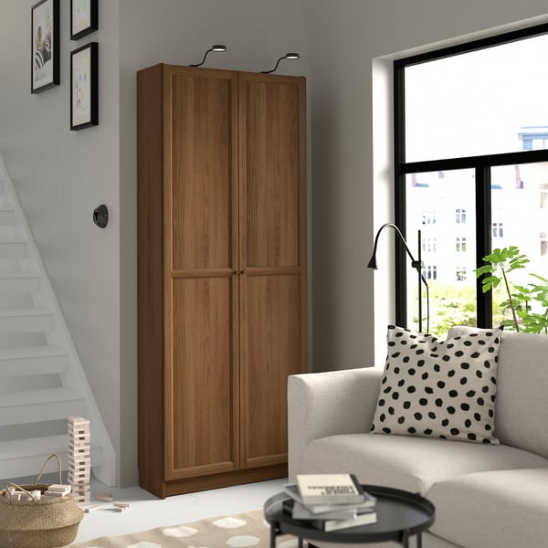 Wooden BILLY bookcase next to stairs & cosy chair, tall with double doors.