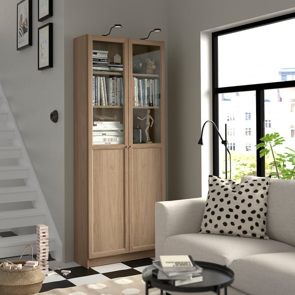 Living room with light brown BILLY bookcase, books, decor, staircase, white couch, and coffee table with plant.