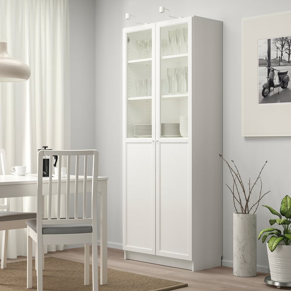 High white BILLY cabinet with glass doors, holding plates, next to small plant, grey chair by dining table.