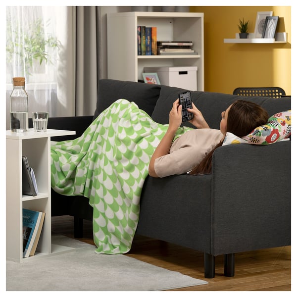 Person on sofa with blanket, using phone. Beside it, laptop and water on shelf, yellow wall, bookshelf.