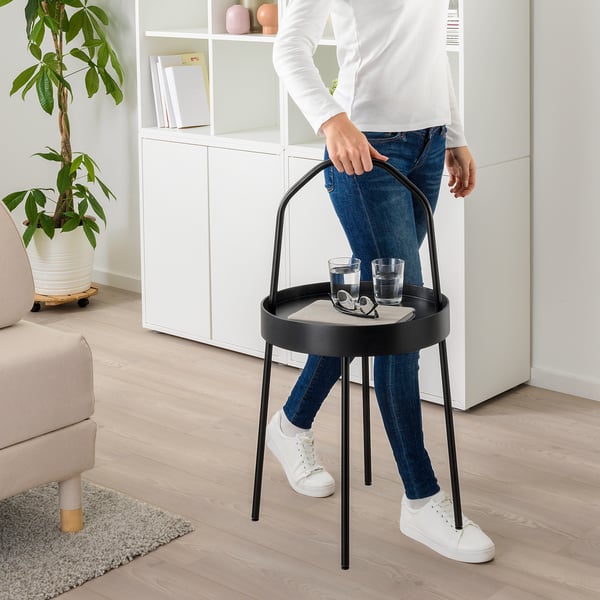 Person holding portable, round side table with handle. Table holds two glasses and coasters. Table has black metal frame and light-colored top.