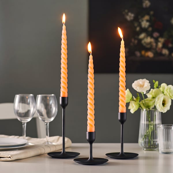 Three twisted orange candles in black holders light a dinner table, near glasses and white flowers in a vase.