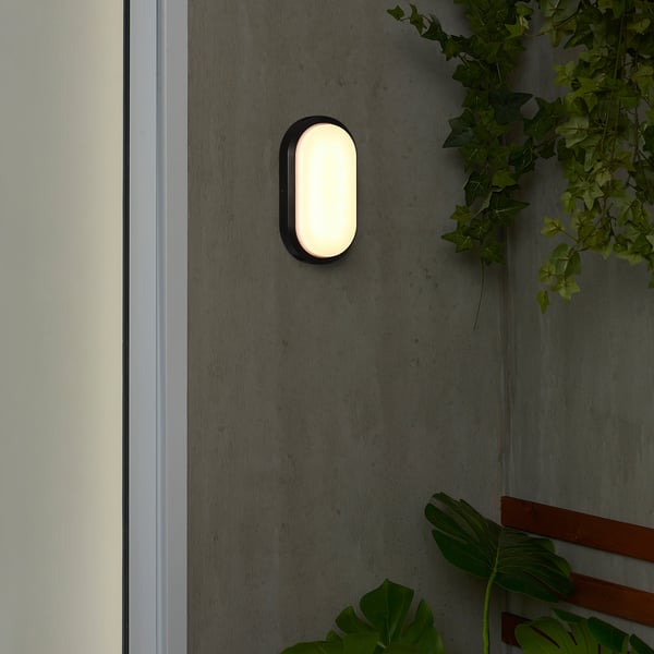 Small, oval, black wall light, DÄCKSBÅT, with a polycarbonate shade, emits warm light, mounted on a grey wall, beside green plants.