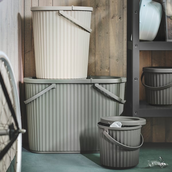 Stack of three gray striped DAMMÄNG bins, largest at base, smallest on top, with foldable handles and lids, positioned against a rustic wall.