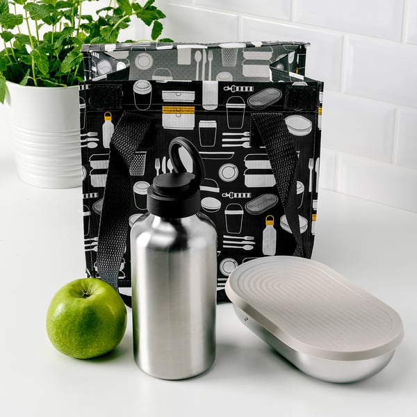 Black patterned bag with kitchen utensils, silver water bottle, green apple, and open metal container on white counter.