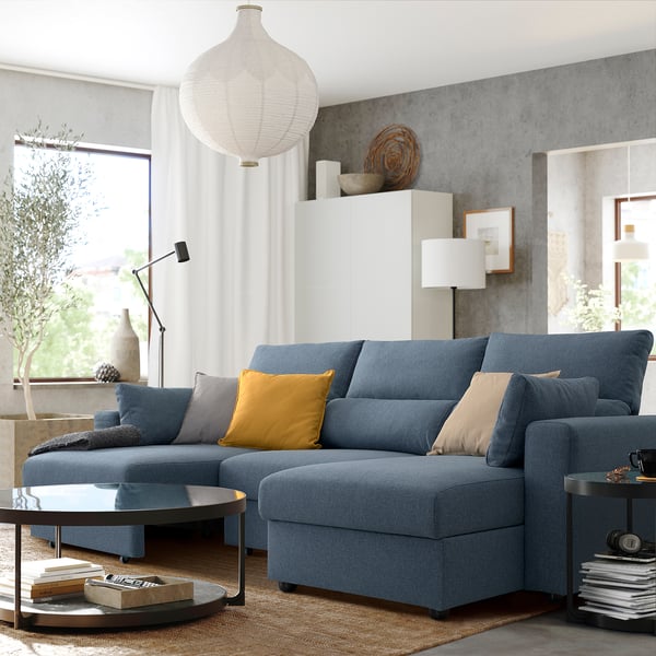 Modern living room with ESKILSTUNA blue sectional, large cushions, yellow pillow, sleek coffee table, side lamp, large rug, and natural decor.