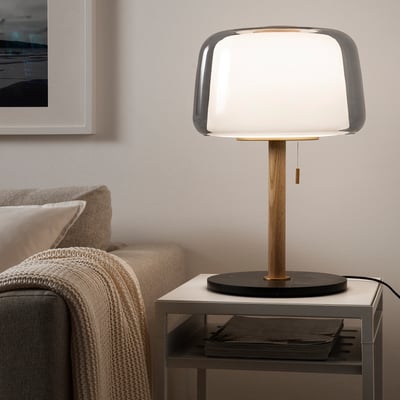 Modern EVEDAL lamp on table, glass shade & wooden stand, near cosy grey sofa, stylish & balance design, fits well in living space.