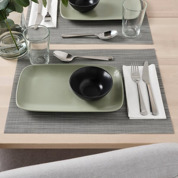 A dining setup with FÄRGKLAR bowl on rectangular green plate, cutlery, and glass on grey placemat.