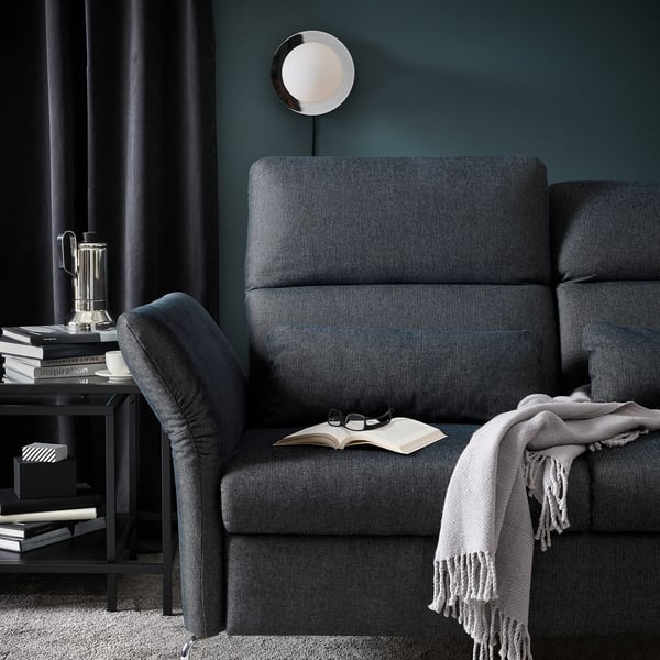 Cosy living room with grey FAMMARP sofa, dark teal walls, round wall light, side table with books and coffee press, blanket on sofa, dark curtains.