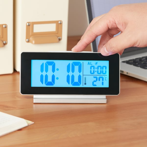 A hand interacts with a modern FILMIS led clock on a wooden desk. The digital display shows time, alarm, and temperature.