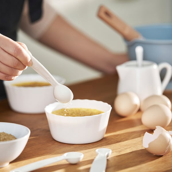 Hand pours liquid into small white bowl on wooden table, surrounded by eggs, measuring spoons, and kitchen utensils.