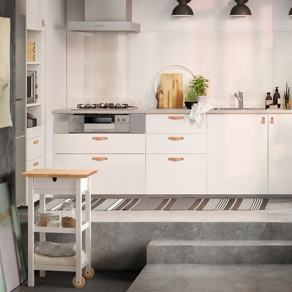 Small white wooden kitchen trolley, holding jars, next to large white cabinetry, black stove, and concrete countertops, under hanging black lamps.