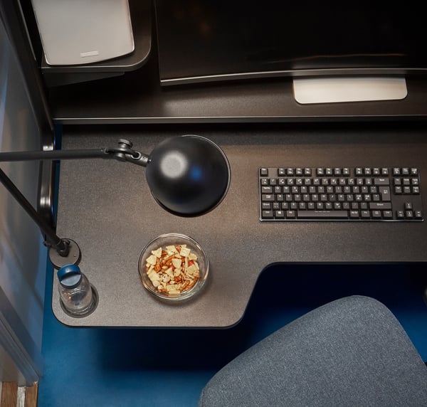 A modern black desk setup featuring a monitor, keyboard, lamp, and snack bowl.