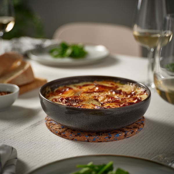 Small dark-grey stoneware bowl sits on cork coaster, filled with layered dish. Surrounded by plates and wine glass, on a light tablecloth.