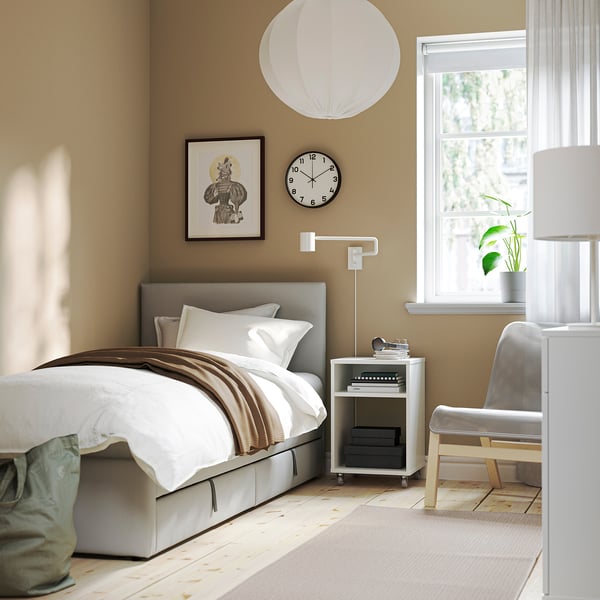 Grey GLADSTAD bed with white linens, brown throw, padded headboard, bedside lamp, small chair, and window with white curtains in a minimalist bedroom.