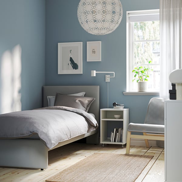 Modern bedroom with grey bed, blue walls, pendant light, sheer curtains, bedside table with books, plant, wooden chair, artwork, wall light.