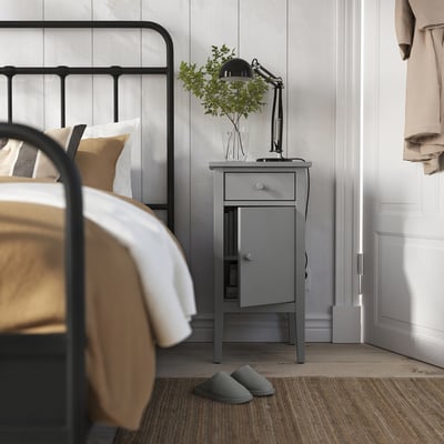 Bedroom scene with black metal bed, grey GULLABERG bedside table, lamp, plant, and slippers.