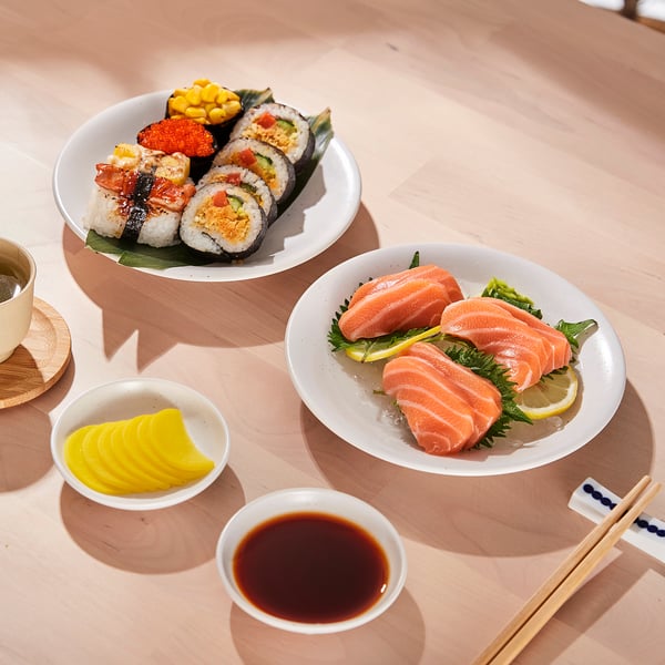 Table setting with sushi rolls, raw salmon, pickles, soy sauce, and chopsticks on white bowls and plates.