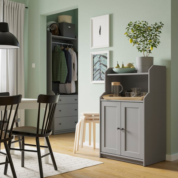 Modern living space with green walls, featuring HAUGA grey cabinet with wooden top, black chairs, white stools, lemon tree, and paintings.