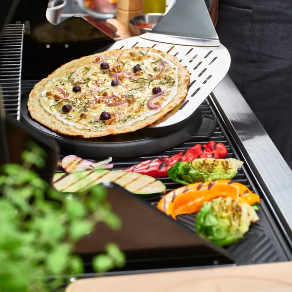 Pizza on cast iron pan on grill, veggies grilling around, handles for lifting.