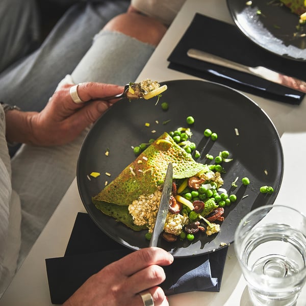 Person holding fork above dish, featuring green crepes and peas. The dish is on a black plate, with a knife and glass beside it.