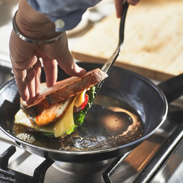 A hand lifts a grilled sandwich with a spatula from a frying pan on a stovetop, showcasing the pans even heat distribution and non-stick surface.