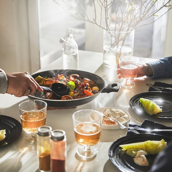 A communal meal is being served from a large non-stick frying pan onto plates. Wine glasses and condiments are also on the table.