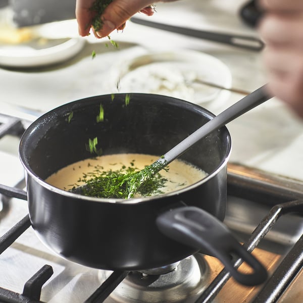 Person sprinkles chopped herbs into a saucepan on a stove. The saucepan has a non-stick coating and is designed for even heat distribution.