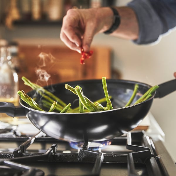 In a hand uses tongs to stir green beans in a wok on a stove. The wok has a glass lid and non-stick coating. Steam rises, indicating cooking.