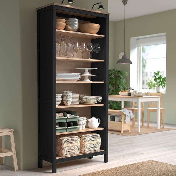 High HEMNES black & wood bookshelf, holding dishes & glasses, beside dining table.