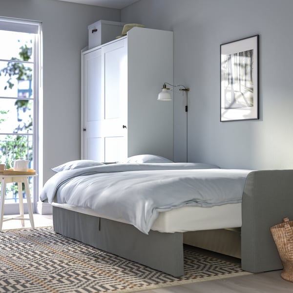 Modern bedroom with light blue walls, a white bed with grey bed skirt, wooden side table, patterned rug, and window with plants.
