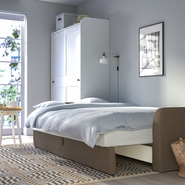 Bedroom with white bed, grey bedframe, wall lamp, tall white cabinet, patterned rug, and wall art.